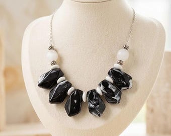 May include: A statement necklace featuring large, irregularly shaped black and white marbled beads. The necklace is strung on a silver chain and accented with white spherical beads. The necklace is displayed on a cream-colored bust.