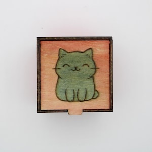 May include: A small, square wooden box with a sliding lid. The top of the box features a green cartoon cat illustration on a light orange background. The box is made of light-colored wood with a dark brown border.