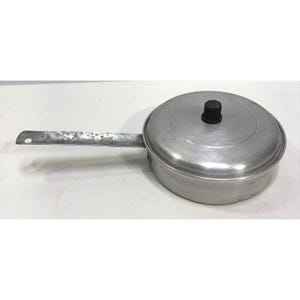 Può includere: Una padella rotonda in alluminio color argento con coperchio abbinato e un lungo manico in metallo piatto e usurato. Il coperchio presenta un piccolo pomello cilindrico scuro al centro. La padella ha una finitura metallica liscia e vissuta.