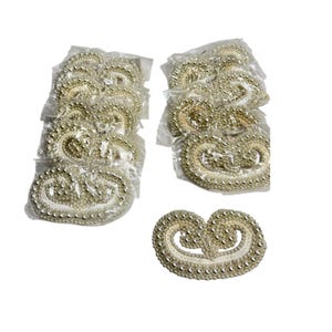 May include: A collection of eleven oval-shaped decorative patches featuring intricate cream-colored beadwork and pearl-like accents. Ten of these patches are individually sealed in clear plastic bags, while one patch is displayed outside of its packaging.