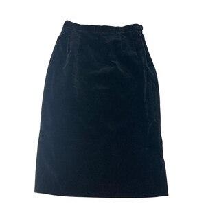 Pode incluir: Uma saia de veludo preta com uma silhueta reta. A saia parece ser até o joelho e tem uma textura macia e lisa. O tecido reflete a luz, dando-lhe um brilho sutil. A saia é de cor sólida e tem um design clássico.