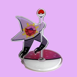 May include: A purple and red acrylic figure of a character holding a staff with a red and white ball on top. The figure is on a clear base with a pink and white design. The background is a solid light purple.