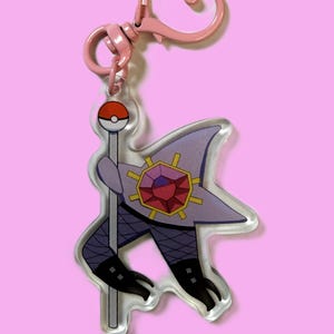 May include: A pink keychain featuring a cartoon character design. The character has a purple star-shaped headpiece with a red gem, black boots, and a staff topped with a red and white ball. The keychain has a pink heart-shaped clasp.