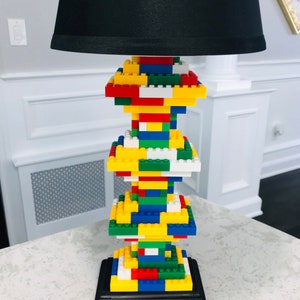 Op de afbeelding: Een zwarte lampenkap met een basis gemaakt van kleurrijke Lego-stenen. De basis is gestapeld in een torenvorm en staat op een wit aanrecht.