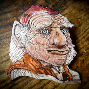 Puede incluir: Una escultura de cuero pintada a mano de un goblin con un sombrero rojo, cabello blanco y ojos azules. El goblin tiene una nariz grande y la boca ligeramente abierta.