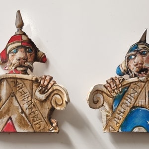 Puede incluir: Dos esculturas de madera tallada de figuras con rostros expresivos y tocados ornamentados. Una figura lleva un casco rojo y blanco, la otra un casco azul y blanco. Ambas figuras sostienen una estructura de madera decorativa.