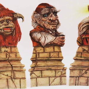 Puede incluir: Tres figuritas coloridas y pintadas a mano de criaturas míticas. Las figuritas están montadas sobre bases de ladrillo con enredaderas que crecen por los lados. Las figuritas son una criatura roja y peluda con cuernos, un hombre de piel verde y barbudo con un sombrero rojo y una criatura marrón y peluda con un sombrero azul.