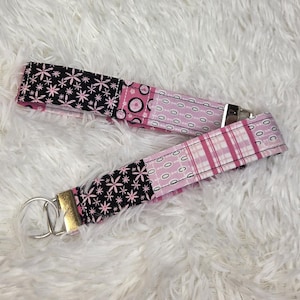 Pode incluir: Um porta-chaves de tecido com um design de patchwork composto por tecido preto com estrelas cor-de-rosa, tecido cor-de-rosa com círculos pretos e tecido cor-de-rosa aos quadrados. A fita é fixada com um grampo metálico prateado e um anel.