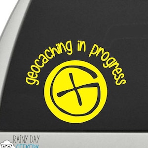 May include: Yellow car sticker with the text "geocaching in progress" and a yellow compass symbol.
