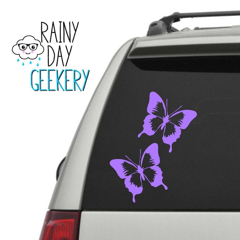 May include: A purple butterfly decal on a car window. The decal features two butterflies in flight, with their wings spread wide. The butterflies are a light purple color and have a simple, elegant design.