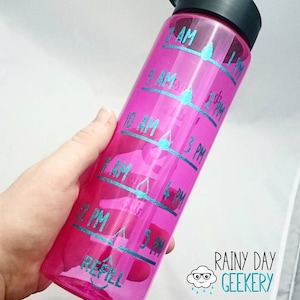 Puede incluir: Botella de agua rosa con una escala marcada por horas y una etiqueta de recarga azul. La botella tiene una tapa negra y un icono de gota de agua azul junto a cada marcador de hora. El texto en la botella dice "8 AM", "9 AM", "10 AM", "11 AM", "12 PM", "1 PM", "2 PM", "3 PM", "4 PM", "5 PM", y "RECARGAR".