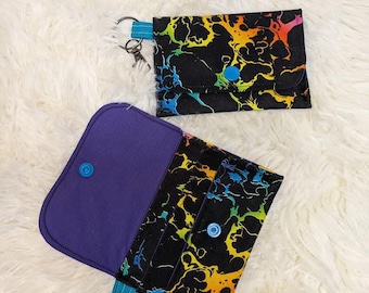 Peut inclure: Deux petits portefeuilles en tissu avec un motif de tourbillon arc-en-ciel et un fond noir. Les deux portefeuilles ont une fermeture à bouton bleu et une sangle turquoise avec un porte-clés argenté.