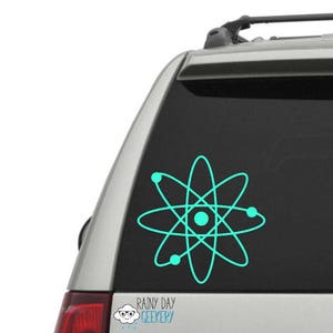 May include: A teal blue atom symbol decal on a silver car. The decal is a simple design with a circle in the center and five orbiting electrons.