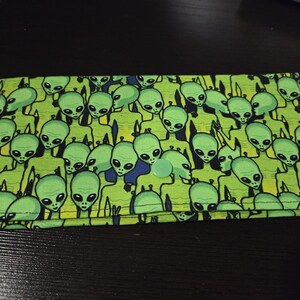 May include: A green and blue wallet with a repeating pattern of green alien figures. The wallet is closed with a light green button. The background is a dark surface.