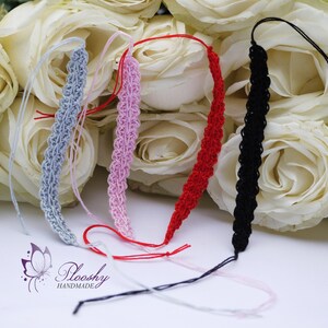 May include: Four crocheted bracelets in different colours: grey, pink, red, and black. The bracelets are laid out on a white surface with white roses in the background. The text "Plooshzy HANDMADE" is visible in the bottom centre of the image.