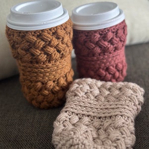 May include: Three crocheted coffee cup cozies in brown, pink, and beige. The cozies are made with a textured stitch pattern.