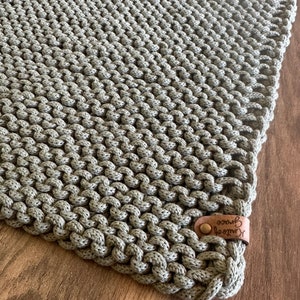 Może przedstawiać: Ręcznie robiony, szałwiowy zielony koc z makramy o teksturowanym, węzłowym wzorze. Koc posiada sk&oacute;rzaną metkę z napisem "Knits of Grace" i guzikiem. Koc jest prezentowany na drewnianej powierzchni.