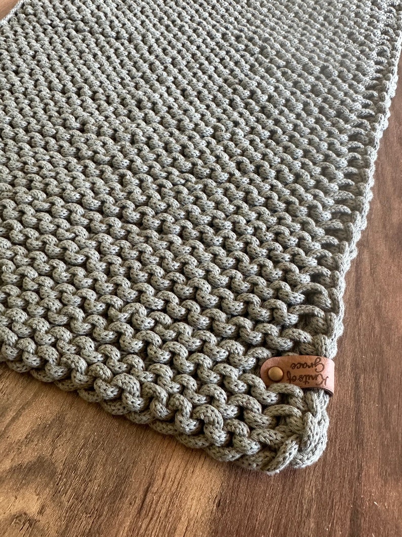 Może przedstawiać: Ręcznie robiony, szałwiowy zielony koc z makramy o teksturowanym, węzłowym wzorze. Koc posiada sk&oacute;rzaną metkę z napisem "Knits of Grace" i guzikiem. Koc jest prezentowany na drewnianej powierzchni.