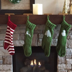 Op de afbeelding: Zes gebreide kerstsokken in rood, groen en wit hangen boven een open haard. De sokken hebben patronen zoals strepen, een rendier, een boom en een sneeuwvlok. De open haard is gemaakt van steen.