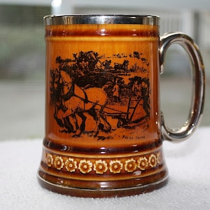 May include: A brown ceramic mug with a silver rim and handle. The mug features a black and white illustration of horses pulling a plow. The text "Fresh Teams" is printed on the mug.
