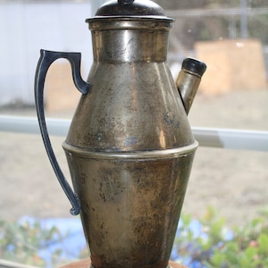 May include: A silver pitcher with a black handle and spout. The pitcher has a rounded body and a flared base. It has a tarnished finish.