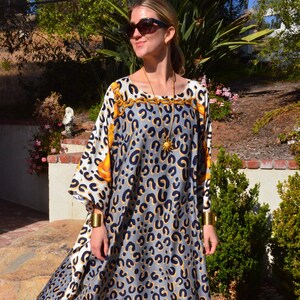 May include: A woman wearing a long, flowing, gray and white dress with a leopard print pattern. The dress has a gold chain trim and a gold sun pendant necklace. She is wearing gold bracelets and sandals.