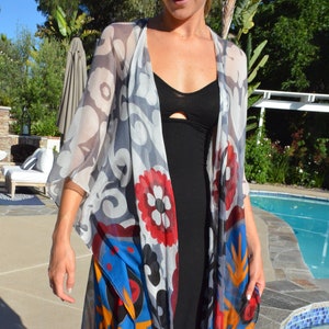 Op de afbeelding: Een doorschijnende, lange, kleurrijke kimono met een zwarte badpak eronder. De kimono heeft een blauw, rood, geel en wit bloemenpatroon. De persoon staat bij een zwembad.