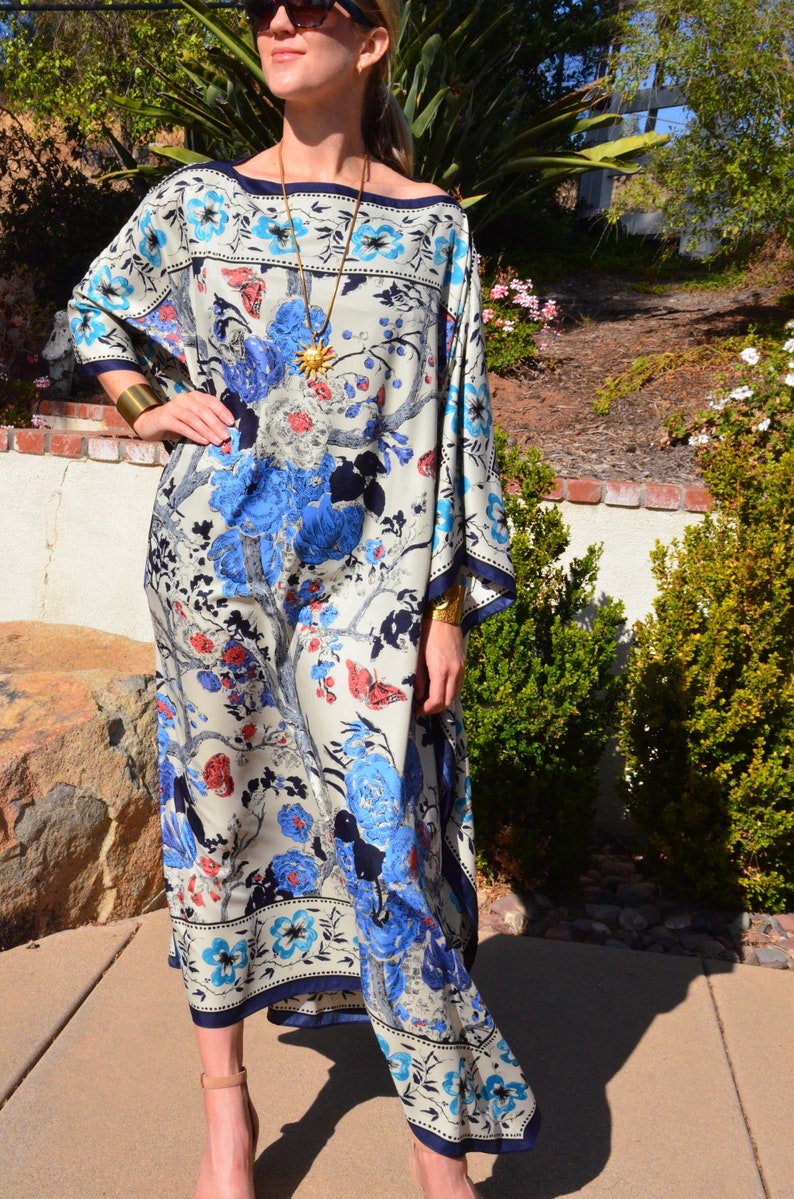 K&ouml;nnte beinhalten: Eine wei&szlig;e, blaue und rote Kaftan mit floralem Muster, einer goldenen Halskette und einem goldenen Armband. Die Kaftan hat eine lockere Passform und einen langen, flie&szlig;enden Rock.