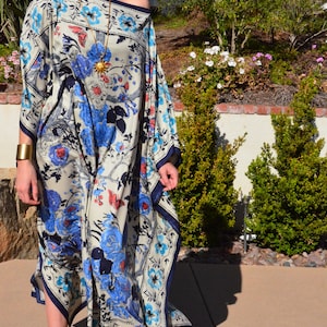 K&ouml;nnte beinhalten: Eine Frau tr&auml;gt ein langes, flie&szlig;endes, schulterfreies Kleid mit einem Blumendruck in Blau-, Wei&szlig;- und Rott&ouml;nen. Das Kleid hat einen dunkelblauen Saum und eine goldene Sonnen-Anh&auml;ngerkette.