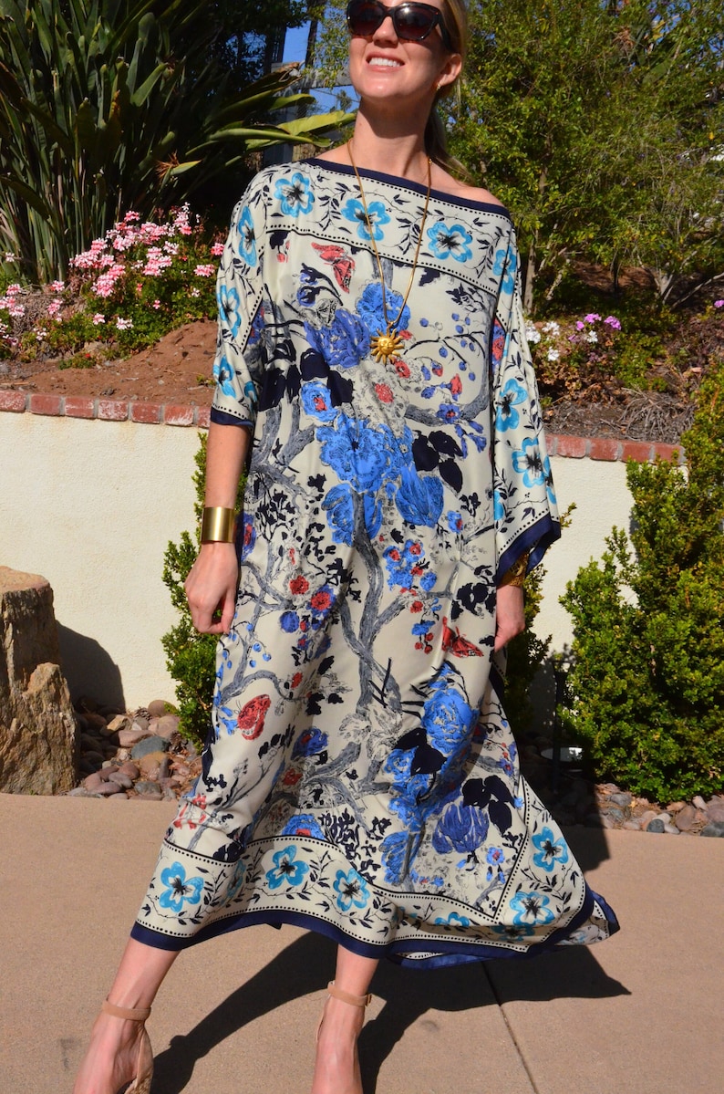 K&ouml;nnte beinhalten: Eine Frau tr&auml;gt ein wei&szlig;es, blaues und rotes Maxikleid mit floralem Muster, eine goldene Halskette und ein Armband. Das Kleid hat eine weite, flie&szlig;ende Silhouette und einen runden Ausschnitt.