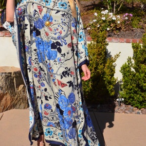 K&ouml;nnte beinhalten: Eine Frau tr&auml;gt eine lange, wei&szlig;e Kaftan mit einem blauen und roten Blumendruck. Die Kaftan hat eine weite, flie&szlig;ende Silhouette und einen tiefen V-Ausschnitt. Die Frau tr&auml;gt eine goldene Halskette und eine Sonnenbrille.