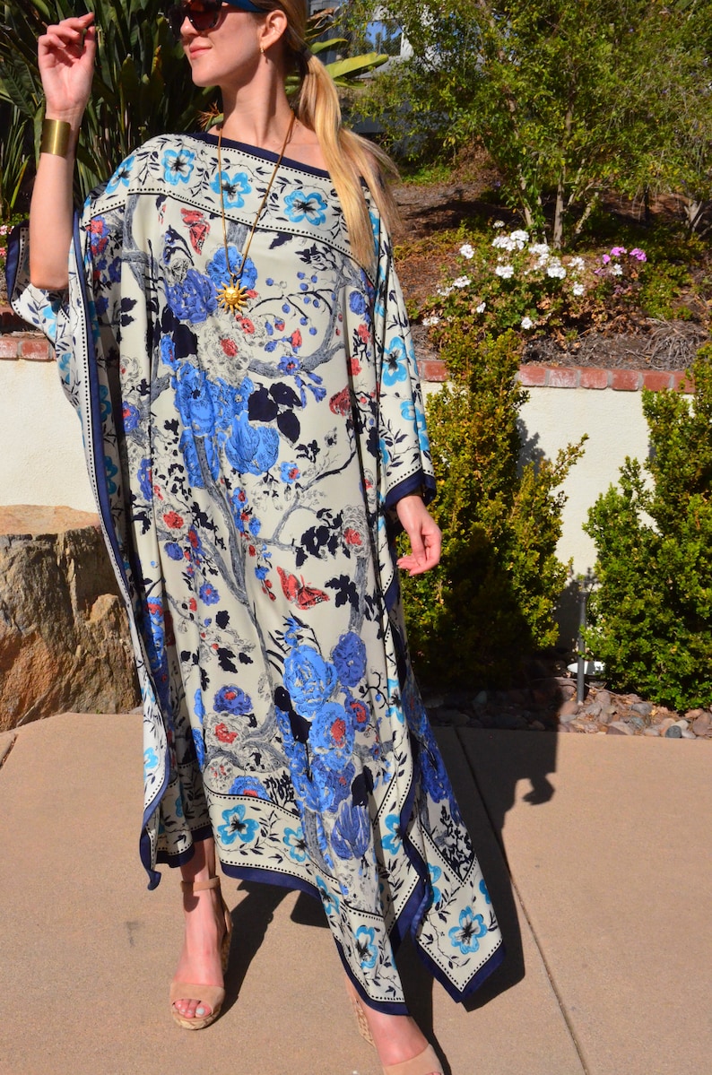 K&ouml;nnte beinhalten: Eine Frau tr&auml;gt eine lange, wei&szlig;e Kaftan mit einem blauen und roten Blumendruck. Die Kaftan hat eine weite, flie&szlig;ende Silhouette und einen tiefen V-Ausschnitt. Die Frau tr&auml;gt eine goldene Halskette und eine Sonnenbrille.
