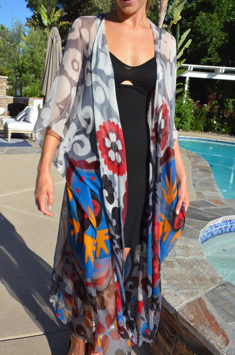 Op de afbeelding: Een doorschijnende, lange, kleurrijke kimono met een zwarte badpak eronder. De kimono heeft een blauw, rood, geel en wit bloemenpatroon. De persoon staat bij een zwembad.