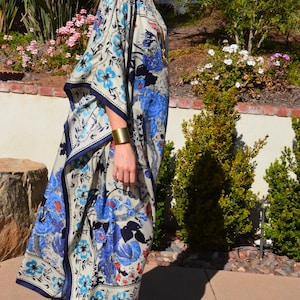 K&ouml;nnte beinhalten: Eine Frau tr&auml;gt eine lange, flie&szlig;ende wei&szlig;e und blaue Kaftan mit floralem Muster und einem goldenen Armband am Handgelenk. Sie steht auf einer Betonterrasse mit gr&uuml;nen Pflanzen im Hintergrund.