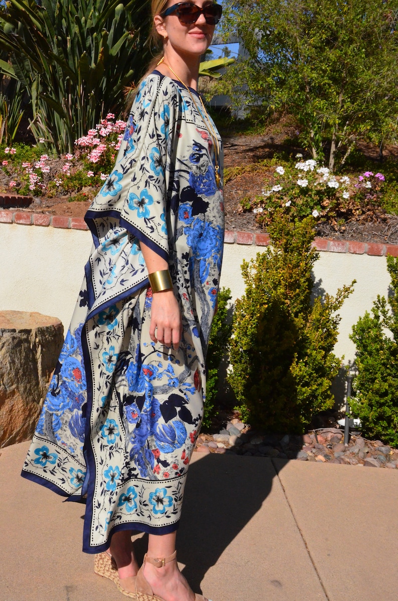 K&ouml;nnte beinhalten: Eine Frau tr&auml;gt eine lange, flie&szlig;ende wei&szlig;e und blaue Kaftan mit floralem Muster und einem goldenen Armband am Handgelenk. Sie steht auf einer Betonterrasse mit gr&uuml;nen Pflanzen im Hintergrund.