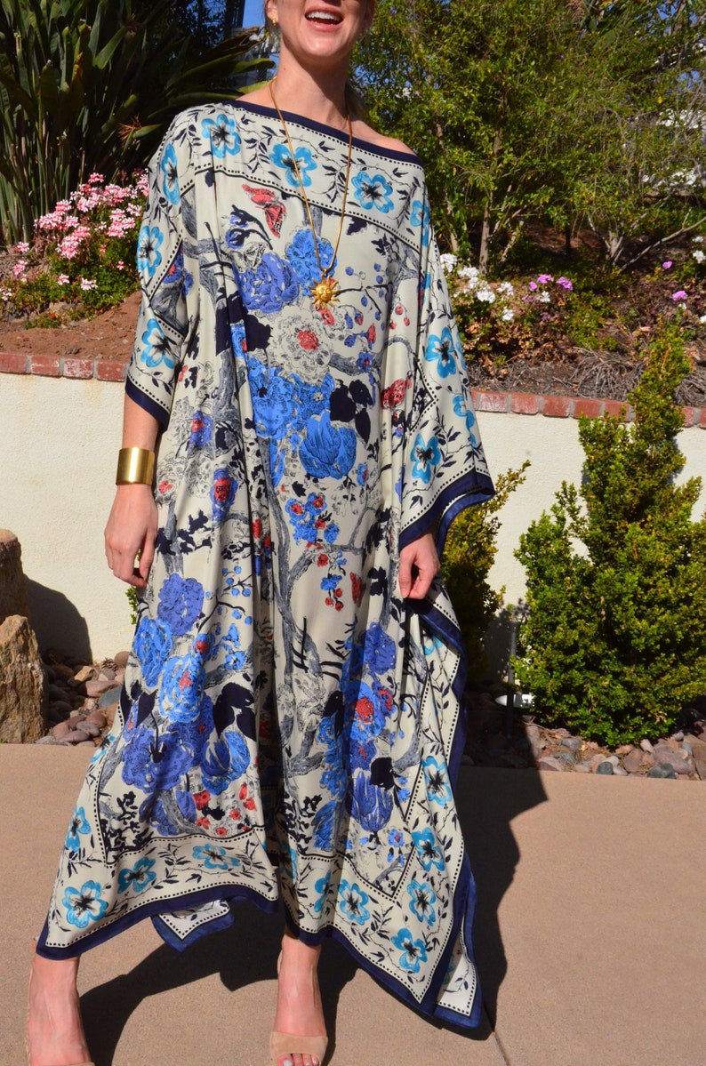 K&ouml;nnte beinhalten: Ein wei&szlig;es Maxikleid mit einem blauen, roten und schwarzen Blumendruck. Das Kleid hat einen quadratischen Ausschnitt und eine flie&szlig;ende Silhouette. Das Kleid ist aus einem leichten Stoff gefertigt und perfekt f&uuml;r warmes Wetter.