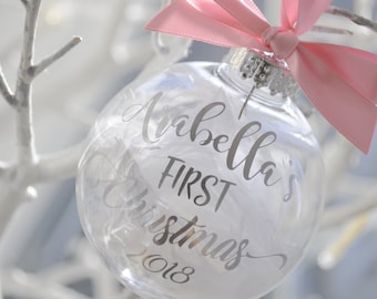 May include: A clear glass Christmas bauble with a pink ribbon and the text "Arabella's First Christmas 2018" in silver lettering.