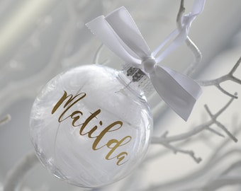 May include: A clear glass bauble filled with white feathers and a gold inscription that reads "Matilda". The bauble is tied with a white satin ribbon.