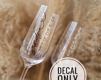 May include: Two clear champagne flutes with black vinyl decals. The flute on the left has the text "Sandra Mother of the Bride". The flute on the right has the text "Mother of the Groom". The text "Decal Only" is printed in a white circle on a brown background.