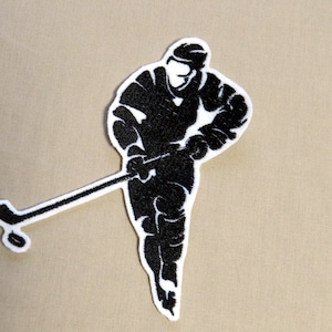 Könnte beinhalten: Schwarz-weiß gestickter Aufnäher eines Hockeyspielers, der mit einem Schläger Schlittschuh läuft. Der Spieler trägt einen Helm und ein Trikot.