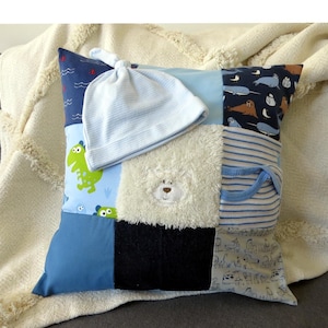 Könnte beinhalten: Ein quadratisches Patchwork-Kissen aus verschiedenen blauen und weißen Stoffquadraten, darunter Muster mit Dinosauriern, Meerestieren und Streifen, mit einem flauschigen weißen Bärengesicht in der Mitte und einer hellblau gestreiften Babymütze darauf.