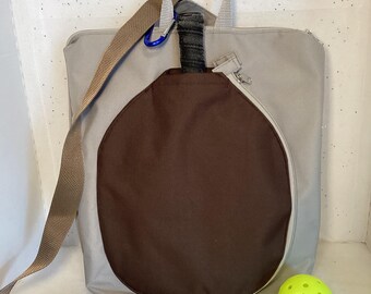 May include: A gray and brown fabric bag with a zippered pocket and a strap. The bag has a large, round pocket on the front for a paddle. A yellow pickleball is in the foreground.