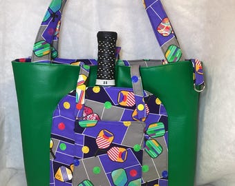 May include: Green tote bag with a pocket featuring a colorful pickleball paddle pattern. The bag has matching straps and a pickleball paddle handle is visible. A yellow pickleball sits in front of the bag.