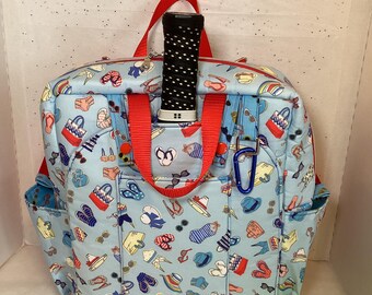 May include: A blue backpack with a summery print of sandals, sunglasses, and beach bags. The backpack has red straps and a black pickleball paddle is sticking out of the top pocket. A yellow pickleball is in the foreground.
