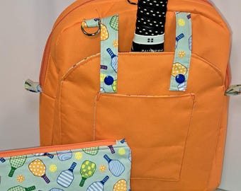 May include: An orange pickleball backpack with a blue strap, a matching zippered pouch, and a yellow pickleball. The backpack has a pocket and a black and white pickleball paddle handle is visible. The pouch has a light blue background with pickleball graphics.