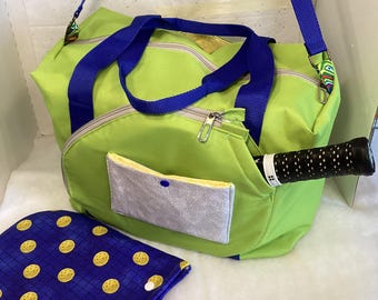 May include: A lime green sports bag with a blue strap and zipper. The bag has a small gray pocket with a blue button. A blue and yellow patterned paddle cover with a black paddle is in the foreground.