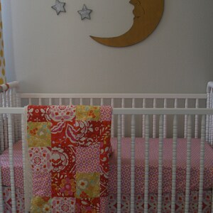 Könnte beinhalten: Ein weißes Babybett mit einer rosa-roten Blumen-Steppdecke und einem rosa-weißen gemusterten Bettrock. Das Babybett steht vor einer weißen Wand mit einem goldenen Halbmond und drei silbernen Sternen.