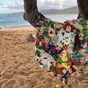 以下が含まれることがあります： 砂浜のビーチで、木の枝にカラフルなパッチワークのキルトが掛けられています。キルトには、熱帯の花柄がプリントされています。