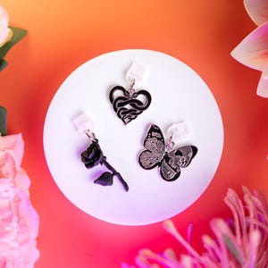 Puede incluir: Tres pendientes negros y plateados con diseños únicos se exhiben sobre una superficie blanca. Los pendientes incluyen un corazón de serpiente, una rosa y una mariposa con diseño de calavera. El fondo presenta arreglos florales rosas y naranjas.