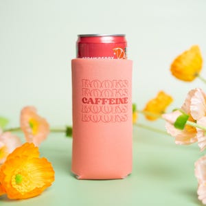 Op de afbeelding: Een koraalkleurige drankisolator met de woorden "BOOKS CAFFEINE BOOKS" in een retro lettertype. Een rood blikje aardbeiensoda zit in de isolator. De achtergrond toont oranje en roze bloemen op een lichtgroen oppervlak.